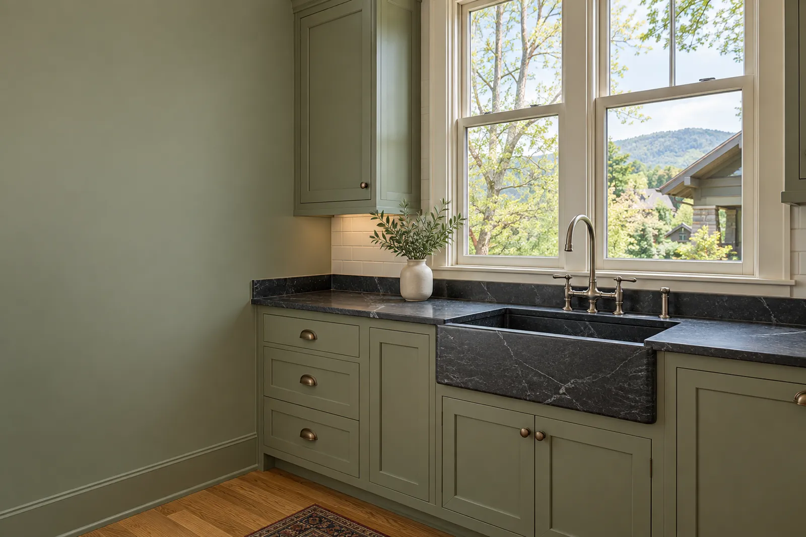 Sage-green shaker cabinets with a dark soapstone counter and a matching apron-front sink, a brass bridge faucet, white subway tile and double-hung windows onto trees and a wooded ridge