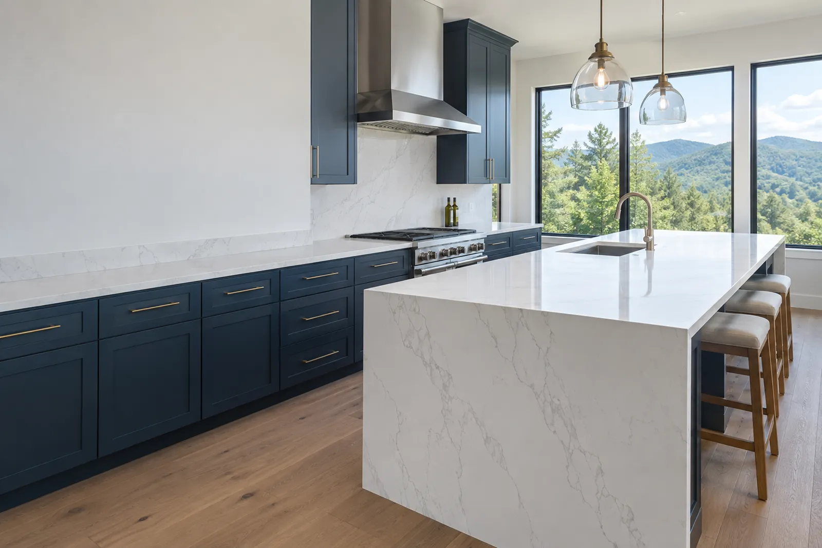 Navy shaker cabinets with white quartz counters and a waterfall-edge quartz island, a stainless hood, glass pendants, wood bar stools and black-framed windows onto green wooded hills