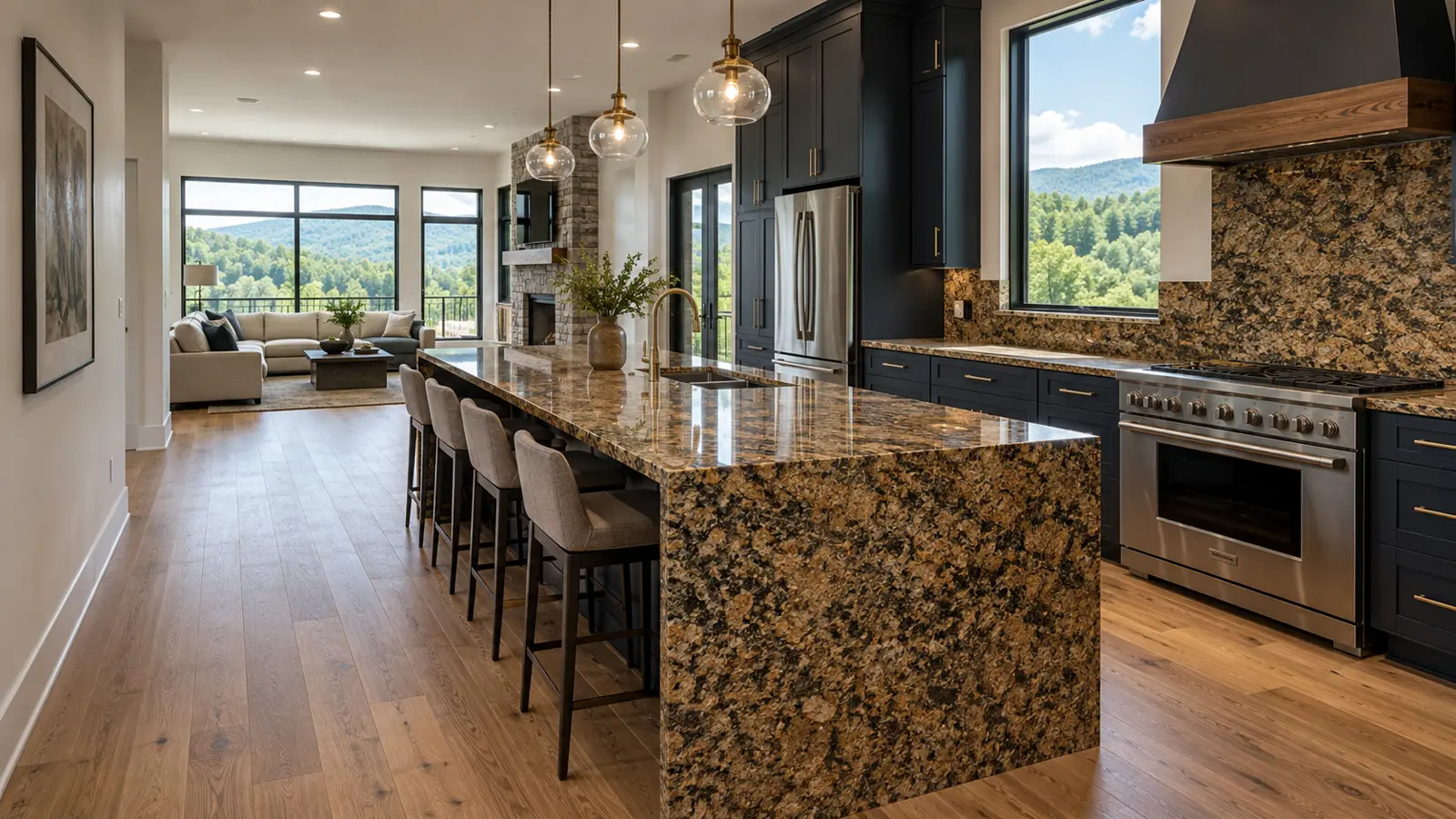 Long open-plan view down a waterfall-edge island in warm brown and gold speckled granite, pendant lights overhead, navy cabinets and a living area with mountain-view windows beyond